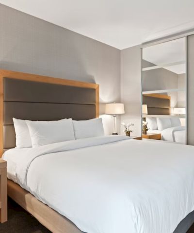 A modern hotel room with a large bed, two nightstands, table lamps, a mirrored wall, and neutral decor.