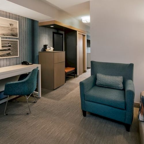 A modern hotel suite: a white desk with a blue chair, a framed photo wall, a teal wall, a tall lamp, a blue armchair, and a cozy bed corner.