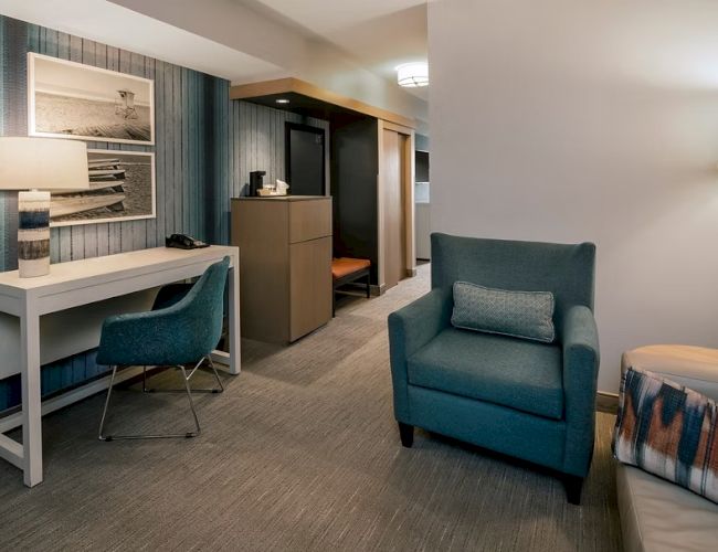 A modern hotel suite: a white desk with a blue chair, a framed photo wall, a teal wall, a tall lamp, a blue armchair, and a cozy bed corner.
