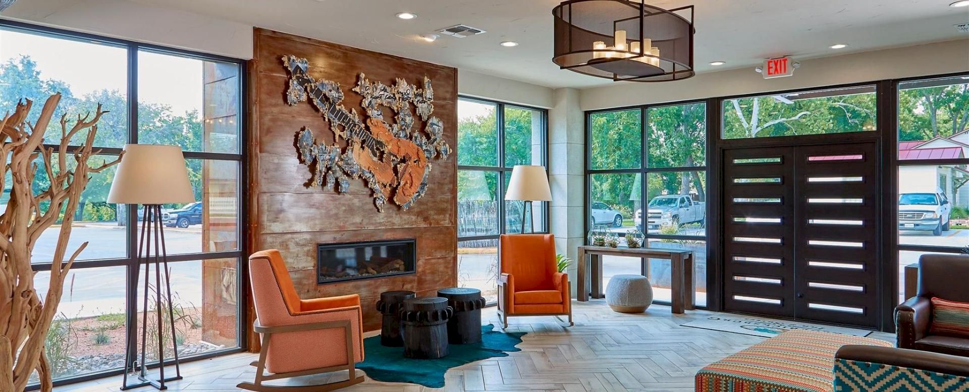 A stylish hotel lobby with a wooden feature wall, a metal animal sculpture above a fireplace, orange chairs, large windows, and modern lighting.