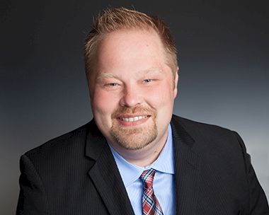 A smiling man in a dark suit, light blue shirt, and striped tie posing for a professional headshot.