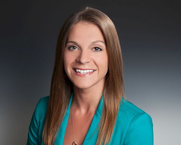 A professional headshot of a smiling woman with long light brown hair, wearing a teal blazer, posing against a neutral backdrop.