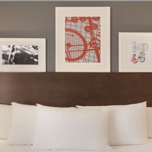 A neatly made hotel bed with white linens, a dark wood headboard, two bedside lamps, and three framed art prints above it.