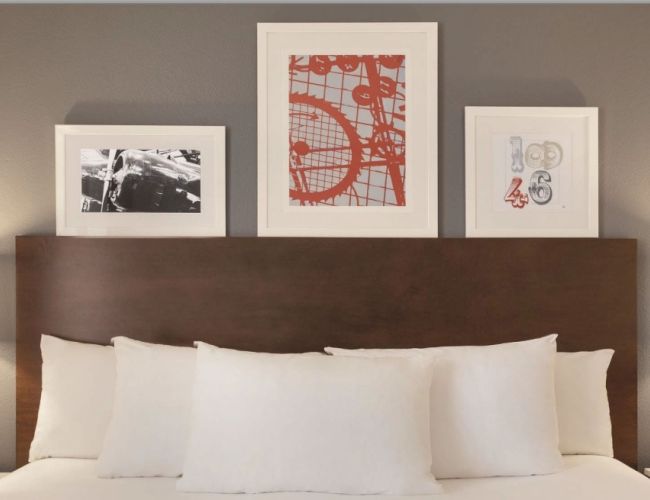 A neatly made hotel bed with white linens, a dark wood headboard, two bedside lamps, and three framed art prints above it.