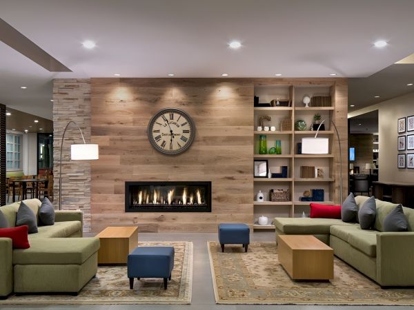 A modern hotel lobby with a central fireplace, big wall clock, wood paneling, and sectional sofas in olive green with colorful cushions.