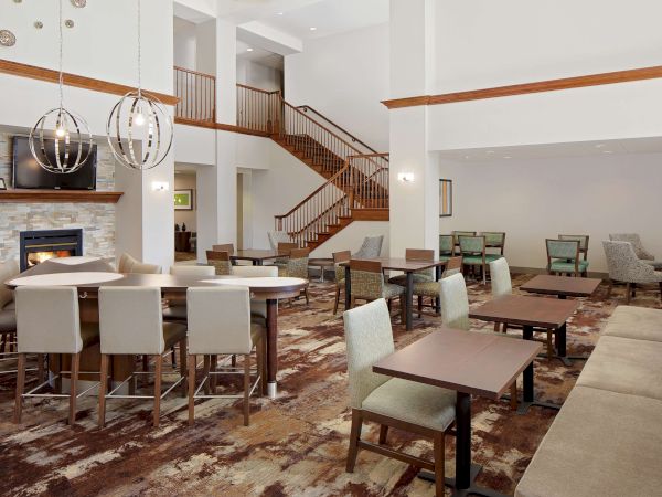 A spacious hotel lobby with a bar, dining area, a grand staircase, and lounge seating, all in warm, neutral tones and modern decor.