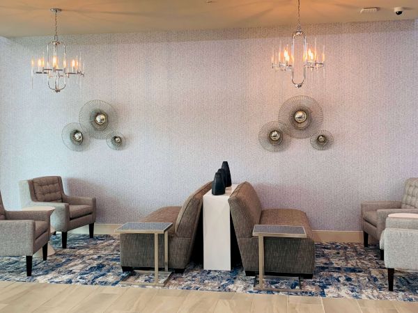 A modern seating lounge with light chairs, a central table, and decorative wall lamps; patterned blue rug underfoot and soft ambient lighting. End.