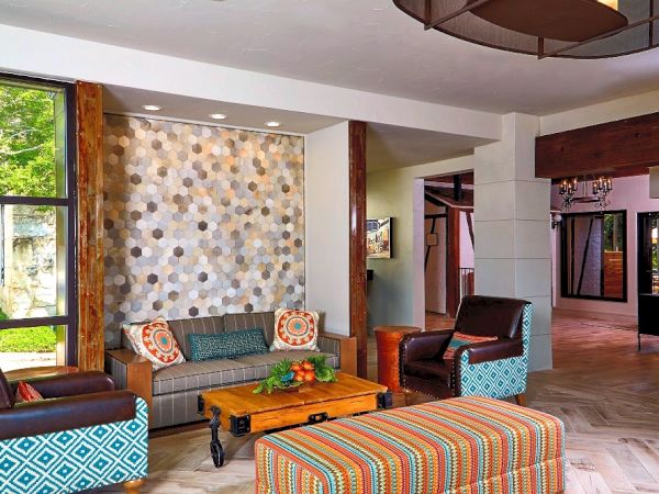A bright, cozy living room with colorful striped sofa, patterned armchairs, a wooden coffee table, tiles on the feature wall, and large windows.
