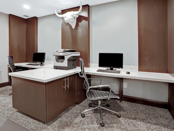 A modern, wood-toned office with a central island desk, a printer, two computer stations, a swivel chair, and a mounted bull skull decor above.
