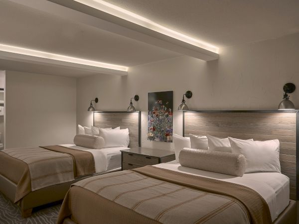 Two double beds in a modern hotel room with neutral tones, bedside lamps, and a decorative artwork between the beds.