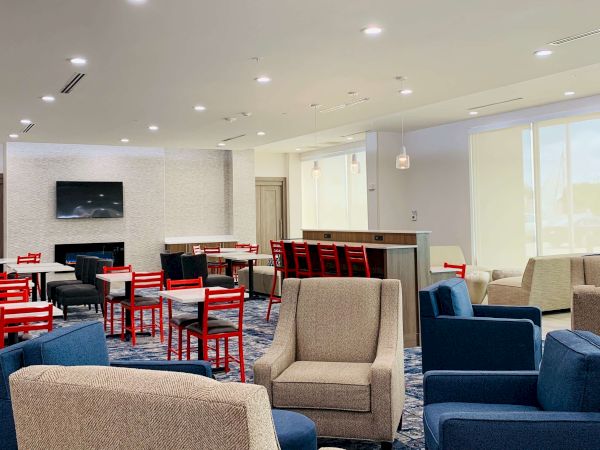 A modern lounge area with blue and beige armchairs, red dining chairs, a small bar, a TV on the white brick wall, and large windows, bright and inviting.