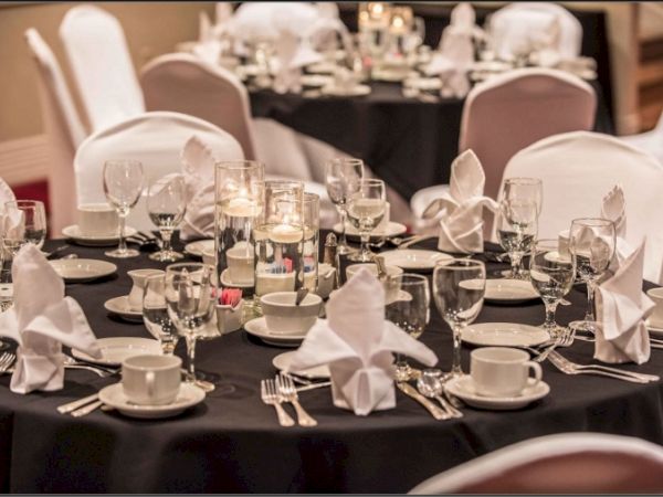 A formal dining setup with multiple round tables, white napkins folded, glassware, plates, and silverware arranged for a banquet.