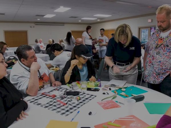 A diverse group collaborates around a sunlit table, drawing, chatting, and sharing papers and colorful crafts in a workshop setting.