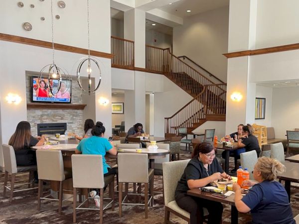 A hotel lobby/restaurant with people dining at tables, a TV on the wall, a modern staircase, and a bright, airy open space.