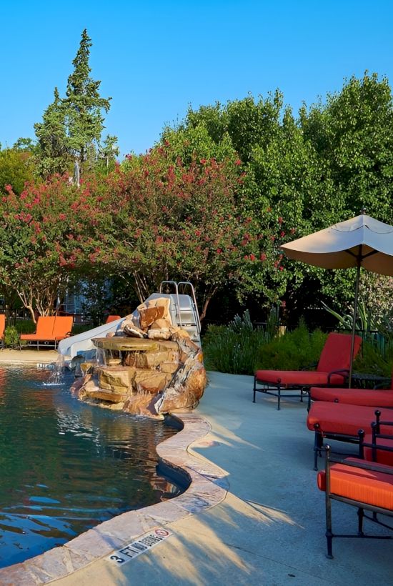 A private backyard pool with stone accents, lounge chairs, a waterfall feature, lush trees, and outdoor seating under sunny skies, all inviting relaxation.