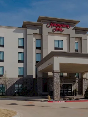 A Hilton Garden Inn-style hotel with a beige facade, a porte-cochère entry, and a red sign that reads “Hampton Inn” in the center, under a clear sky.