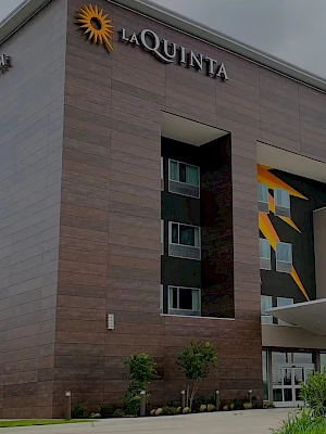 A modern La Quinta hotel exterior with a tall brick facade, glass entrance, and a prominent sign/crest, landscaping in front.