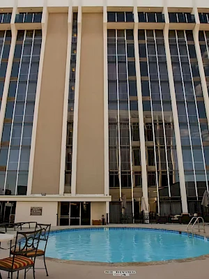 A tall hotel-style building with a circular pool in the foreground, glass windows, and outdoor seating, sunny day by the courtyard.