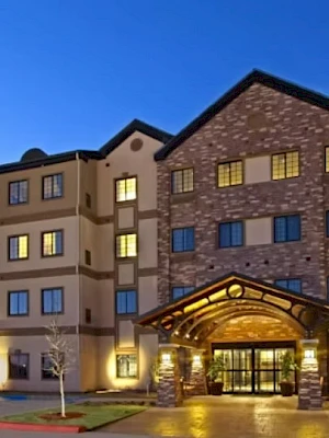 A modern hotel or apartment building at dusk with warm lights, stone accents, and a welcoming entrance canopy. The sky is clear blue.