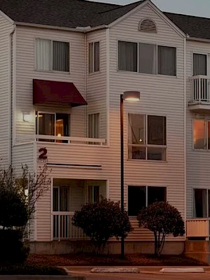 An exterior view of a beige apartment building at dusk, with multiple windows, balconies, and a small landscaped entryway.