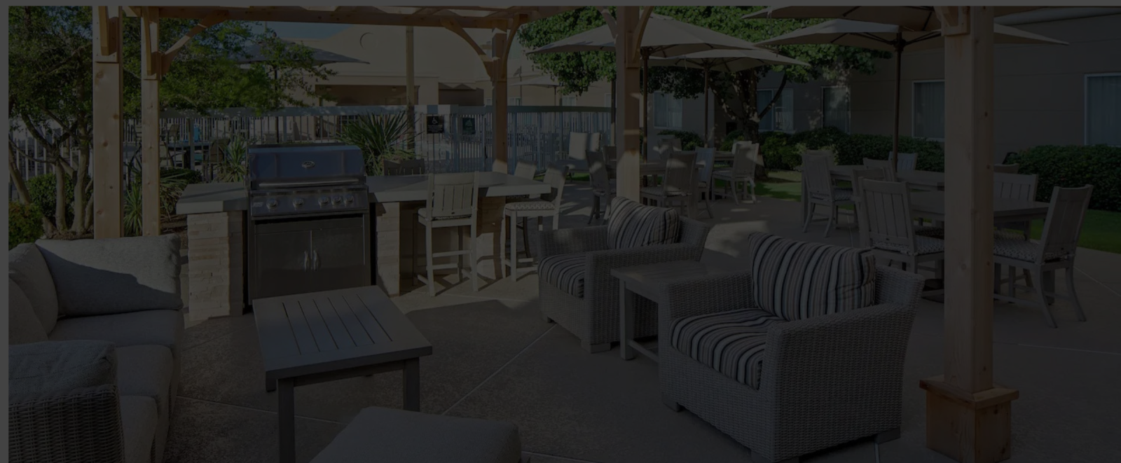 A shaded outdoor seating area with wicker sofas, matching chairs, and a grill, under wooden pergolas with tables and umbrellas, backyard lounge space.