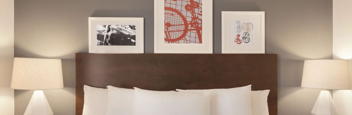 A tidy hotel bedroom with a large wooden headboard, white bedding, two matching lamps, and three framed art pieces above the bed, creating a calm, modern look.