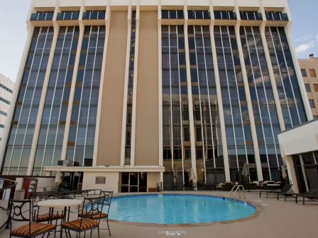 A tall glass-front hotel building with a circular blue pool in the foreground and outdoor seating, white umbrellas, and lounge chairs.
