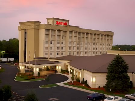 A beige multi-story hotel with a red &ldquo;M&rdquo; sign on top, connected lower wings, a curved entrance, surrounding parking, and trees at dusk.