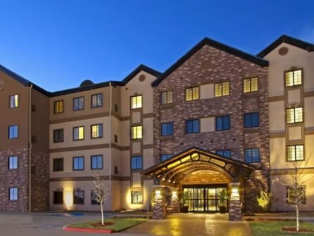 A modern four-story hotel or apartment complex with stone and beige siding and a glass entry, lit up at dusk.