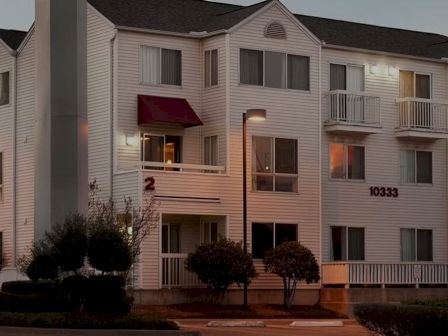 A small white apartment building with two stories, red awnings, numbered exterior doors, and manicured bushes at dusk.