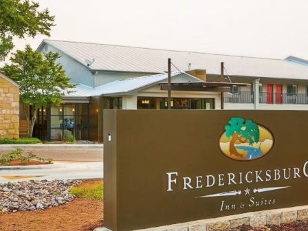 A sign for Fredericksburg Inn & Suites sits near a low stone landscape bed with a 2-story building behind it.